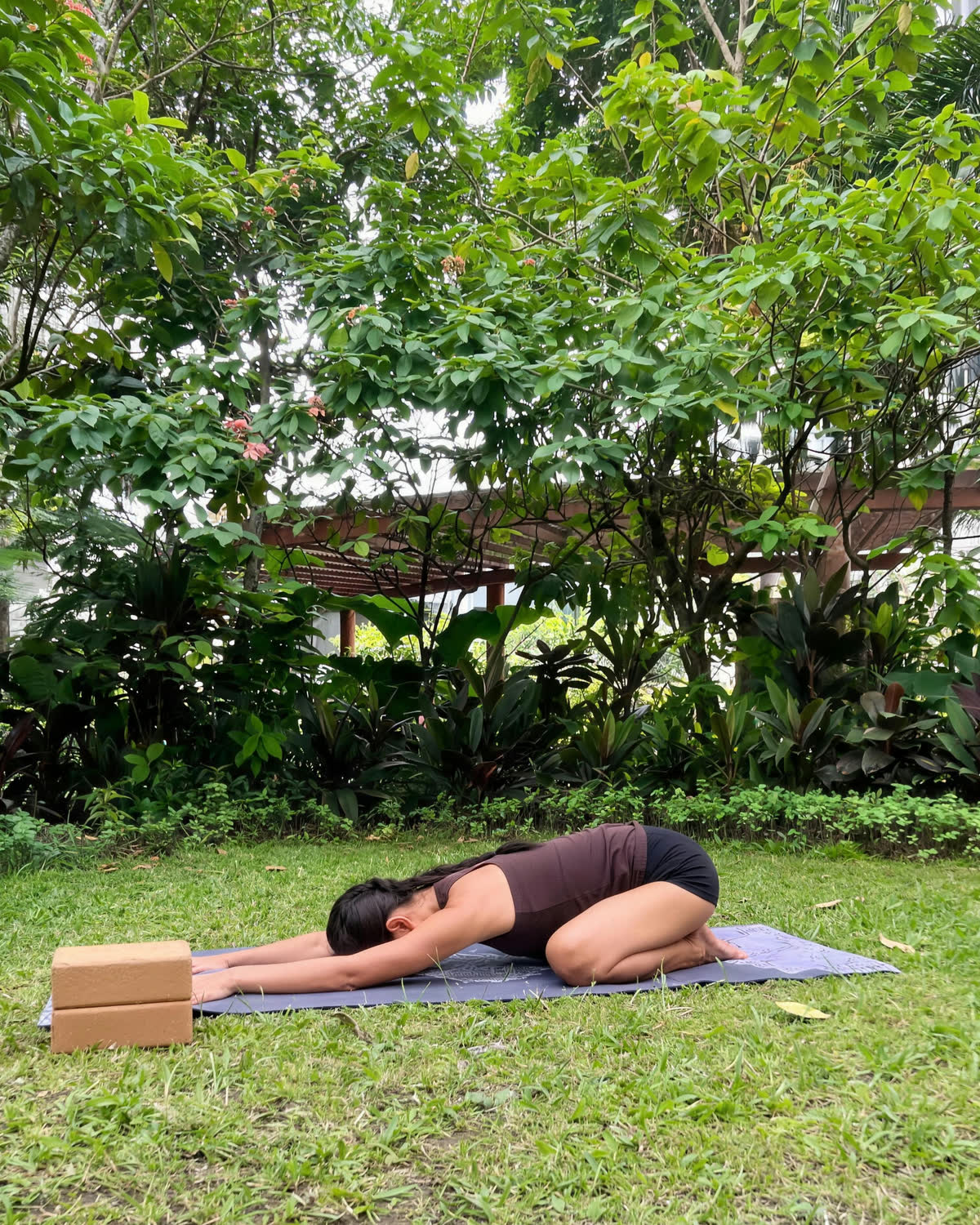 Joahnna Lee Ucol in child's pose on a mat on the grass, in a garden of tropical trees in Metro Manila.