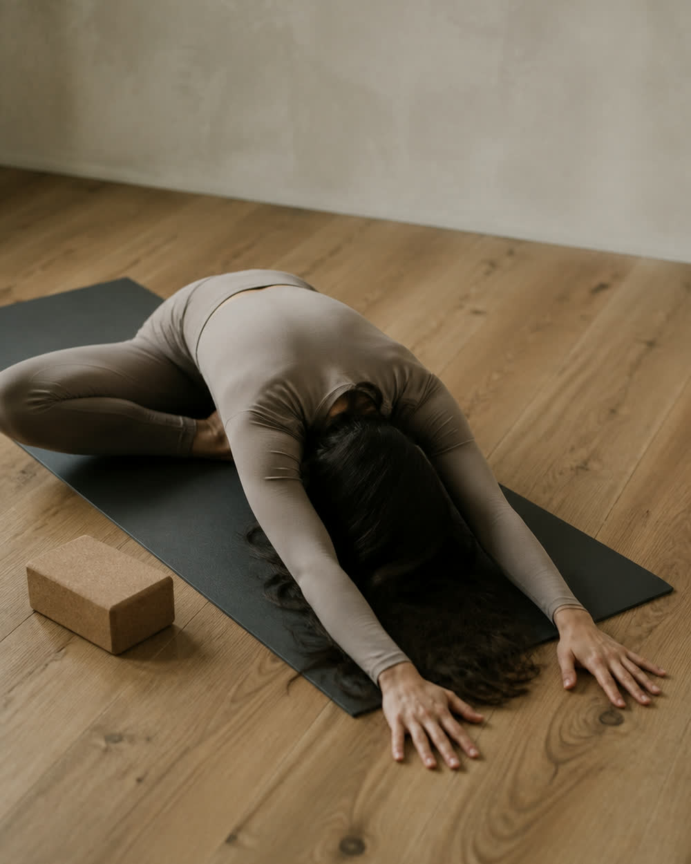 A kneeling forward fold seen from above, arms long on the mat, a cork block resting to one side.