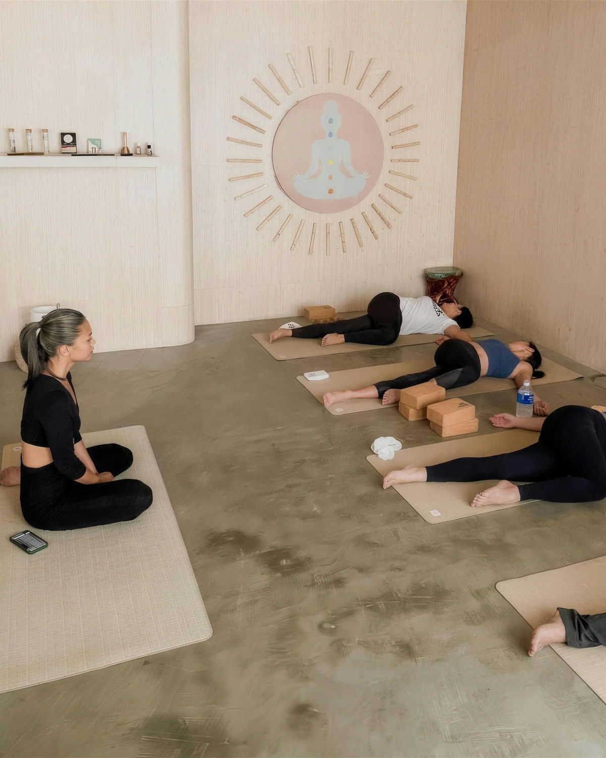 Joahnna kneeling on a mat facing four students lying in a supported twist, in a pale wood and concrete studio.